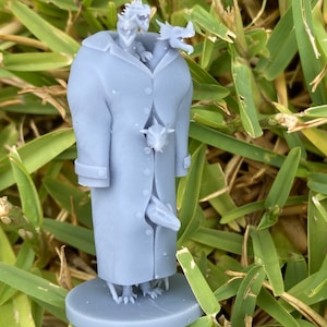 May include: A grey 3D printed miniature of a dragon wearing a long coat. The dragon has four legs and a tail. The coat has a collar and buttons. The miniature is on a round base.
