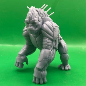 May include: A gray, robotic gorilla figurine in a crouching position. The figure has detailed armor plating and a stern facial expression. White spikes protrude from the back of the head. The figurine is set against a green background.