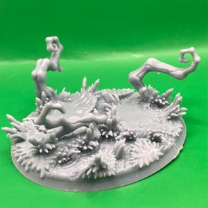 May include: A gray resin miniature base featuring a textured terrain surface with sculpted tufts of grass, small plants, and two curved, leafless tree branches extending upward from the ground. The circular base has a smooth, flat edge.