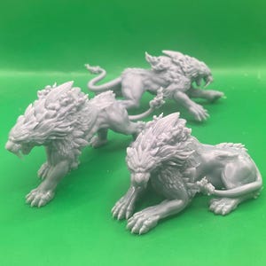 May include: Three unpainted grey resin miniature figurines depicting feline creatures with sharp fangs, muscular builds, and textured manes. Each creature is posed in a dynamic, predatory stance with detailed fur and tail features visible.