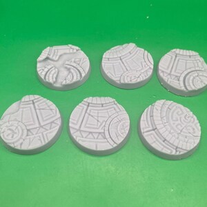 May include: A set of five circular light grey resin bases for miniature figures, each featuring a detailed relief pattern of geometric shapes, interlocking lines, and circular motifs that resemble ancient stone tile flooring or ruins.