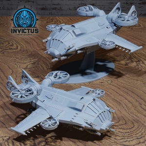 May include: Two gray miniature aircraft models with multiple rotors and wings are displayed on a wooden surface. One model is elevated on a stand. The Invictus Miniatures logo is visible in the upper left corner.