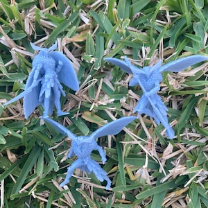 May include: Three light blue miniature figurines of winged creatures, possibly fairies or sprites, are arranged on a bed of green and brown grass. The figurines have detailed wings and are in various poses, suggesting movement or action.