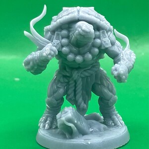May include: A gray, detailed miniature figurine of a humanoid turtle-like creature. The figure has a shell on its back, beaded necklace, and is posed with a weapon. It stands on a circular base, with a rock-like formation at its feet.