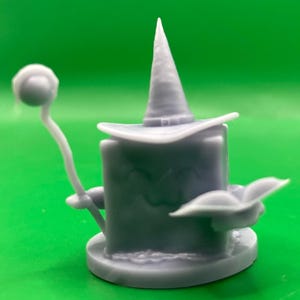 以下が含まれることがあります： 立方体型のキャラクターの3Dプリントフィギュア。とがった帽子をかぶり、杖を持っている。キャラクターは笑顔で、マントを着ている。フィギュアは薄い紫色の塗料で塗られている。