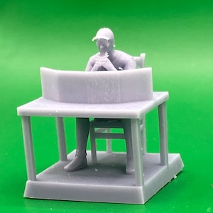 May include: A grey 3D printed figurine of a person sitting at a desk with a curved top. The person is wearing a cap and is looking down at something on the desk.