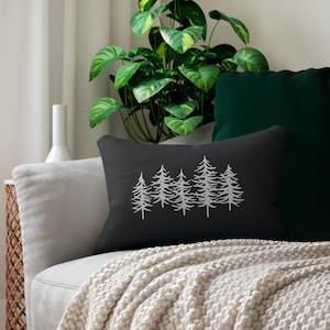 May include: A rectangular throw pillow in a dark charcoal gray color featuring a white line art graphic of a cluster of five evergreen pine trees centered on the front. The pillow has a smooth fabric texture and a simple, clean design.