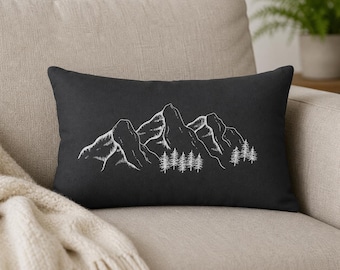 May include: A rectangular throw pillow in a solid dark charcoal gray color. The front of the pillow features a white line art illustration of a mountain range with a cluster of pine trees at the base, rendered in a simple, clean sketch style.