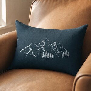 Puede incluir: Un cojín rectangular de color azul oscuro con una ilustración de líneas blancas de una cordillera con un bosque de pinos en la base. El diseño está centrado en la parte frontal del cojín y muestra un boceto detallado de un paisaje.
