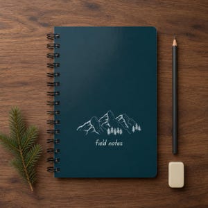 May include: A dark teal spiral-bound notebook featuring a white line drawing of a mountain range with pine trees and the text field notes printed below. A black pencil and a small rectangular eraser are positioned to the right of the notebook.