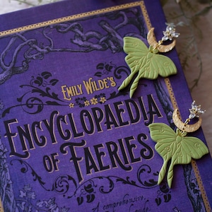 May include: A pair of green and gold earrings featuring a crescent moon and a moth design. The earrings are displayed on a purple book titled "Emily Wilde's Encyclopaedia of Fairies".