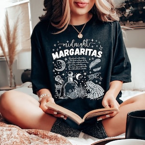 May include: A black t-shirt with a white graphic design that says "midnight margaritas" with a moon, stars, a bottle, a glass, a frog, and feathers.