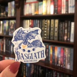 Puede incluir: Una pegatina blanca con una ilustraci&oacute;n azul y negra de un drag&oacute;n volando por el aire. La pegatina tiene el texto "Basgiath War College" en letras azules.