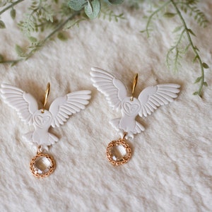 Peut inclure: Une paire de boucles d'oreilles en forme de hibou blanc avec des accents dor&eacute;s et des gouttes de cristal transparent.