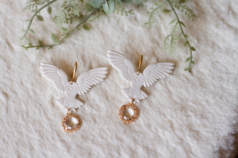Peut inclure: Une paire de boucles d'oreilles en forme de hibou blanc avec des accents dor&eacute;s et des gouttes de cristal transparent.