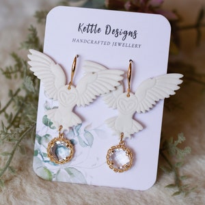 Peut inclure: Boucles d'oreilles pendantes en forme de hibou blanc avec des anneaux dor&eacute;s et des accents de cristal transparent. Les boucles d'oreilles sont pr&eacute;sent&eacute;es sur une carte qui indique "Kettle Designs Handcrafted Jewellery".