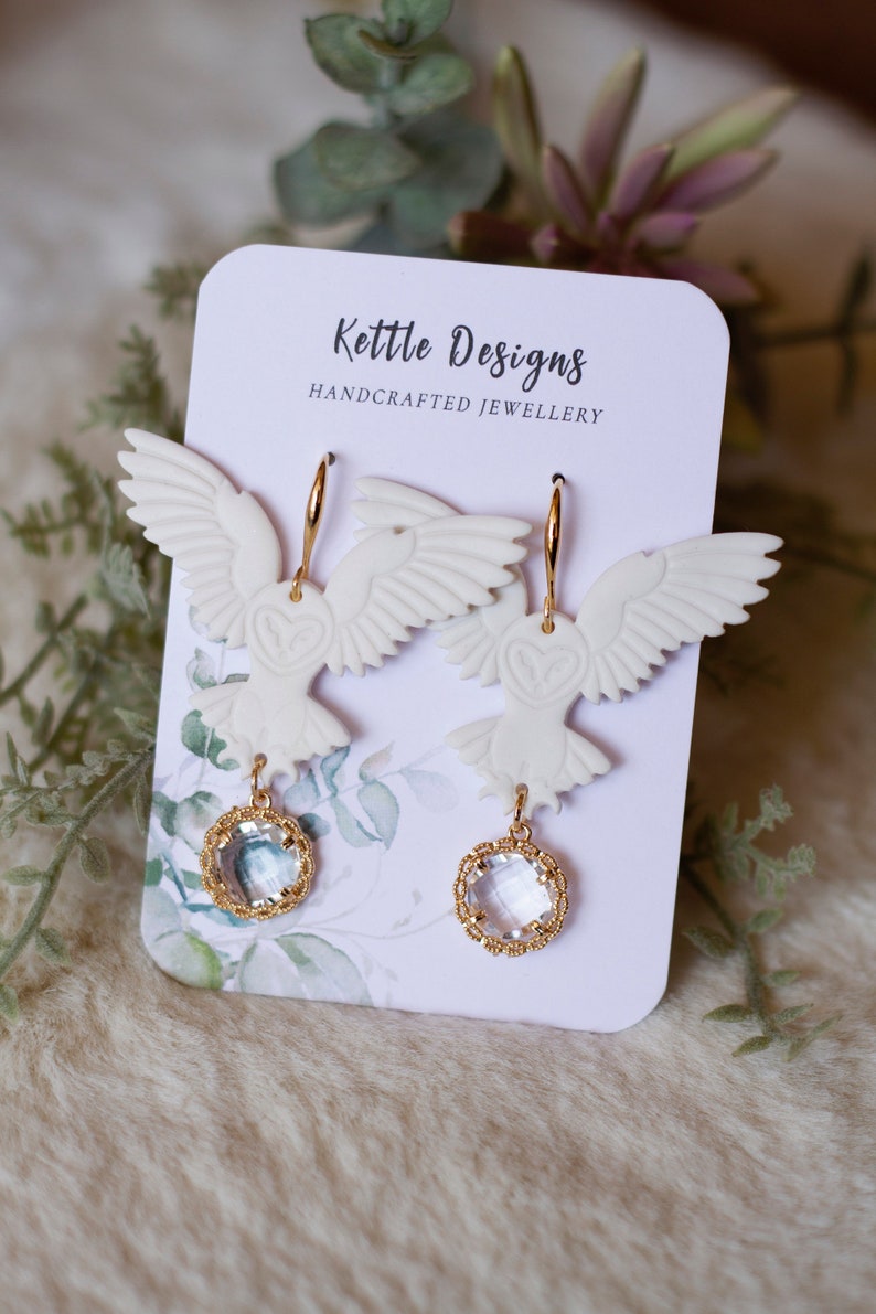 Peut inclure: Boucles d'oreilles pendantes en forme de hibou blanc avec des anneaux dor&eacute;s et des accents de cristal transparent. Les boucles d'oreilles sont pr&eacute;sent&eacute;es sur une carte qui indique "Kettle Designs Handcrafted Jewellery".