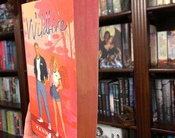 Puede incluir: Un libro de bolsillo titulado "Wildfire" de Hannah Grace, con una portada rosa y naranja que muestra a una pareja de pie frente a un árbol con luces. El libro es parte de la serie Icebreaker.