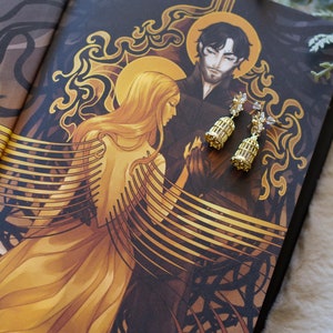 Könnte beinhalten: Ein Paar goldene Käfigohrringe mit klaren Kristall-Akzenten. Die Ohrringe sind auf einem Buchcover abgebildet, das eine Fantasy-Illustration einer Frau mit langen blonden Haaren und einem Mann mit dunklen Haaren zeigt.