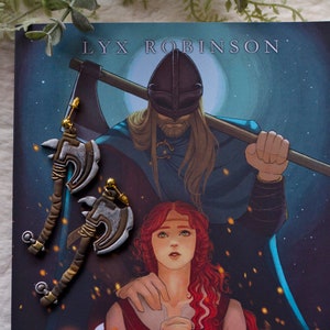 Könnte beinhalten: Ein Paar silberne Axt-Ohrringe mit goldenen Akzenten. Die Ohrringe befinden sich auf einem Buchcover mit einer Fantasy-Illustration eines Mannes und einer Frau. Das Buchcover trägt den Text "LYX ROBINSON".