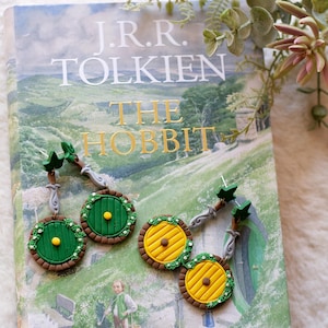 May include: Hobbit door earrings in green and yellow, with a brown border and a yellow doorknob. The earrings are displayed on a copy of "The Hobbit" book by J.R.R. Tolkien.
