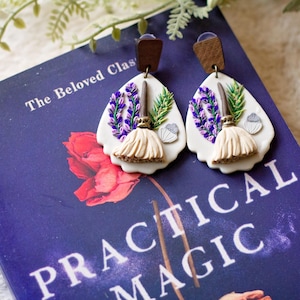 May include: A pair of teardrop-shaped earrings featuring a white base with a raised design of a beige broom, purple lavender sprigs, green pine needles, and a small silver acorn. Each earring is attached to a dark brown wooden rectangular stud top.