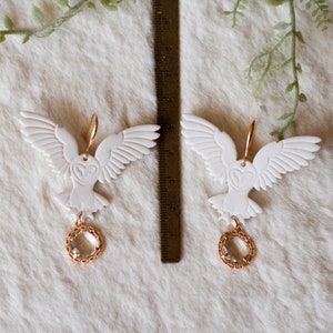 Peut inclure: Une paire de boucles d'oreilles en forme de hibou blanc avec des accents dor&eacute;s et des gouttes de cristal clair. Les hiboux ont les ailes d&eacute;ploy&eacute;es et regardent vers l'avant. Les boucles d'oreilles sont suspendues &agrave; des crochets dor&eacute;s.