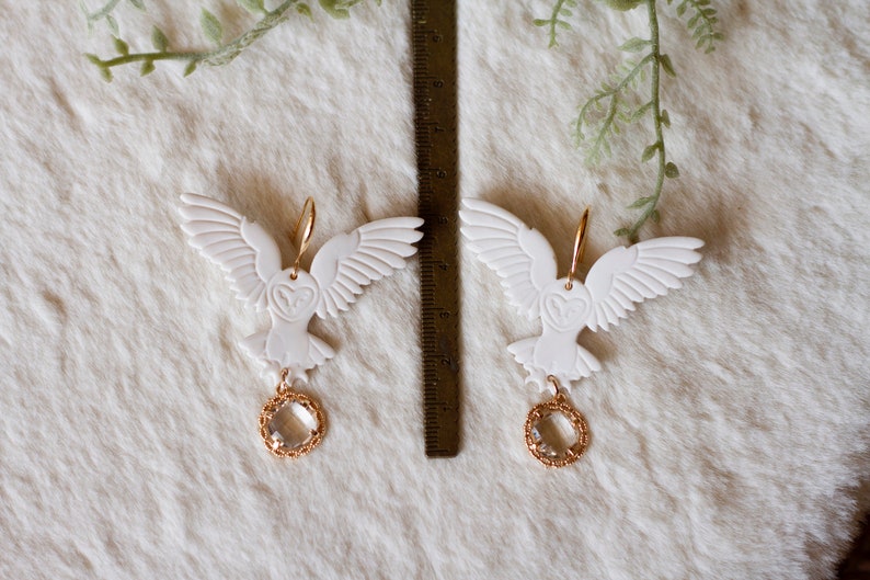Peut inclure: Une paire de boucles d'oreilles en forme de hibou blanc avec des accents dor&eacute;s et des gouttes de cristal clair. Les hiboux ont les ailes d&eacute;ploy&eacute;es et regardent vers l'avant. Les boucles d'oreilles sont suspendues &agrave; des crochets dor&eacute;s.