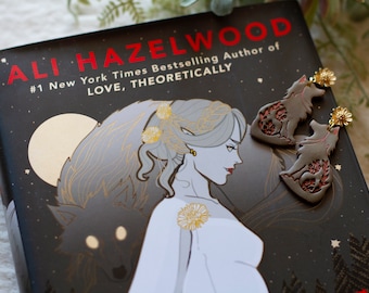 May include: A book cover with a dark background and a woman with long silver hair wearing a white dress. The book title is "ALI HAZELWOOD" and the subtitle is "#1 New York Times Bestselling Author of LOVE, THEORETICALLY". Two wolf-shaped earrings with red and gold accents are on the cover.
