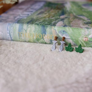 May include: Two pairs of earrings, one pair is silver and shaped like a sword, the other pair is green and shaped like a leaf. The earrings are on a white surface with a book titled "The Hobbit" in the background.
