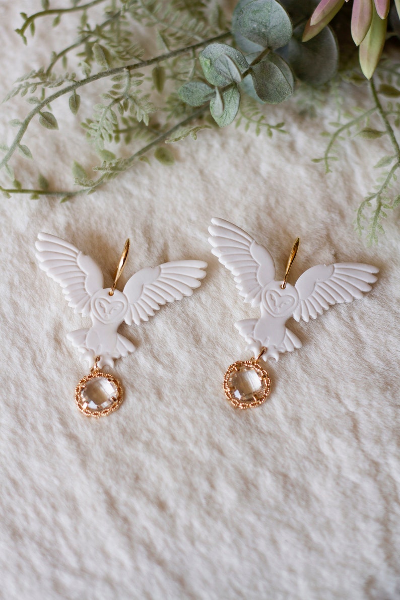 Peut inclure: Une paire de boucles d'oreilles en forme de hibou blanc avec des accents dor&eacute;s et des gouttes de cristal transparent. Les hiboux ont les ailes d&eacute;ploy&eacute;es et sont suspendus &agrave; des cr&eacute;oles dor&eacute;es.