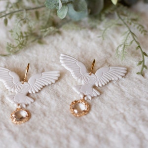 Peut inclure: Une paire de boucles d'oreilles en forme de hibou blanc avec des accents dor&eacute;s et des gouttes de cristal transparent. Les hiboux ont des ailes d&eacute;taill&eacute;es et un motif en forme de c&oelig;ur sur leur poitrine.
