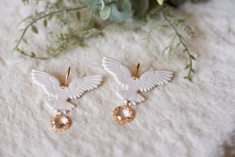 Peut inclure: Une paire de boucles d'oreilles en forme de hibou blanc avec des accents dor&eacute;s et des gouttes de cristal transparent. Les hiboux ont des ailes d&eacute;taill&eacute;es et un motif en forme de c&oelig;ur sur leur poitrine.