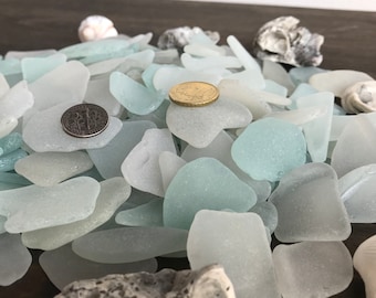 May include: A pile of light blue sea glass, some of which is translucent, with two coins on top. The sea glass is on a dark brown surface.