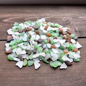 Puede incluir: Un montón de cristales de mar de varios colores, incluyendo blanco, verde, marrón y ámbar. Las piezas son lisas y redondeadas, con un aspecto esmerilado. El cristal de mar está esparcido sobre una superficie de madera marrón oscuro.