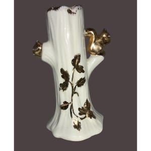 May include: A white ceramic vase shaped like a tree trunk, adorned with gold accents. A gold squirrel sits on a branch, and gold leaves decorate the trunk. The vase has a textured surface and a glossy finish.