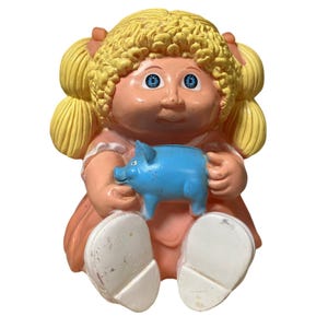 May include: A vintage Cabbage Patch Kids doll figurine, featuring blonde pigtails, blue eyes, and a peach-colored dress. The doll is holding a blue piggy bank. The figurine has white shoes and is sitting down.