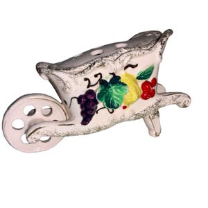 May include: A white ceramic wheelbarrow-shaped planter with a decorative fruit design. The planter features a wheel, handles, and a bed decorated with grapes, pears, and cherries. The edges have a gold trim.