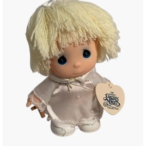 May include: A vintage Precious Moments doll with a light yellow yarn hairstyle and blue eyes. The doll wears a white satin outfit with lace trim and a heart-shaped tag that reads "The Enesco Precious Moments Collection."