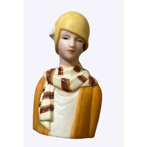 May include: A ceramic figurine bust depicting a person wearing a yellow cloche hat, a white shirt, and a yellow jacket. A scarf with horizontal stripes in shades of brown, red, and white is draped around the neck. The facial features are painted.