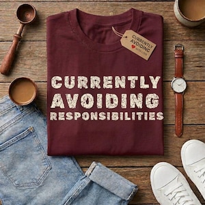 May include: A maroon short-sleeved t-shirt with the white distressed text "CURRENTLY AVOIDING RESPONSIBILITIES" printed on the front. A matching tag attached to the collar displays the same text with a small red heart icon.