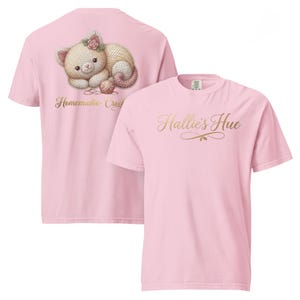May include: A light pink t-shirt featuring a graphic on the back of a crocheted kitten with a pink flower on its head, a ball of yarn, and the text "Homemade Crochet". The front of the shirt displays the gold cursive text "Hallie's Hue".