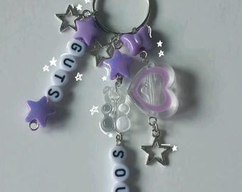 May include: A silver keychain with multiple charms. The charms include purple star-shaped beads, a clear heart-shaped charm, and beads spelling "GUTS" and "SOUR". Additional silver star charms are also attached.
