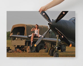 May include: A woman in a white polka dot dress sits on the wing of a black and green airplane. The airplane is parked next to a military truck. The scene is set against a sunset sky.