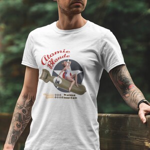 May include: White t-shirt with a graphic of a woman in a red swimsuit sitting on a bomb with the text "Atomic Blonde" and "Warbird Bomber Girls".