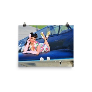 May include: A woman in a floral dress and pink gloves poses on the wing of a blue and white airplane. She is wearing high heels and has her hair styled in a pin-up fashion.