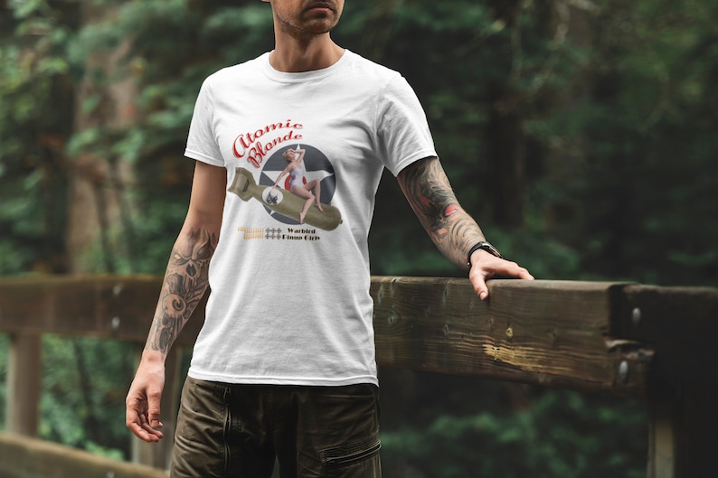 May include: White t-shirt with a graphic of a woman in a red swimsuit sitting on a bomb with the text "Atomic Blonde" and "Warbird Bomber Girls".