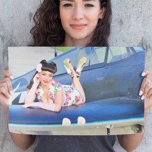 May include: A woman in a floral dress and pink gloves poses on a blue and black airplane wing. She is wearing red heels and has her hair styled in a pin-up style.
