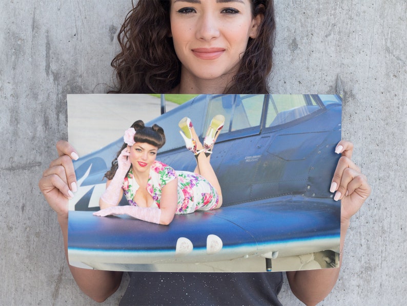 May include: A woman in a floral dress and pink gloves poses on a blue and black airplane wing. She is wearing red heels and has her hair styled in a pin-up style.