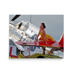 May include: A woman in a yellow and red dress sits on the wing of a silver airplane. The airplane has the number "27" on the side and a star on the tail. The woman is looking to the right and has a red flower in her hair.